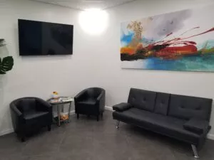 Waiting room at Crystal Bright Smile Dental Office in Tujunga, CA. The room is clean and well-lit, with comfortable black leather chairs and a painting on the wall. A television is mounted on the wall for patients to watch while they wait.