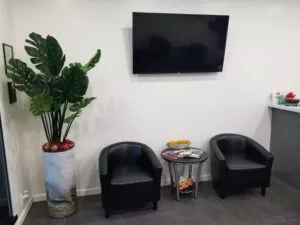 Waiting room with two comfortable black leather chairs with a coffee table between them and a large plant on the left side of one of the chairs with a television mounted on the wall behind them. Waiting room at Crystal Bright Smile Dental Office in Tujunga, CA.
